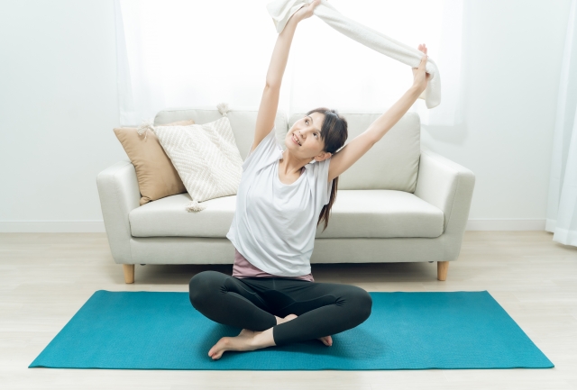 室内で女性が床に座り、タオルを使ってローイング動作を行い、背中を引き締めるトレーニングをしている様子。