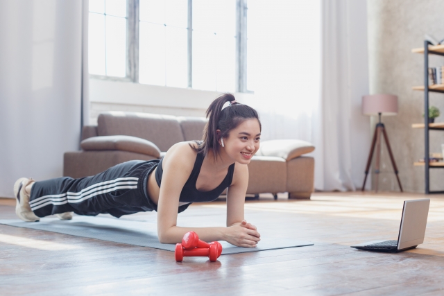 室内でトレーニングウェアを着た女性が、笑顔で椅子プランクを行っている様子。仕事の合間でもできる簡単体幹トレーニング。