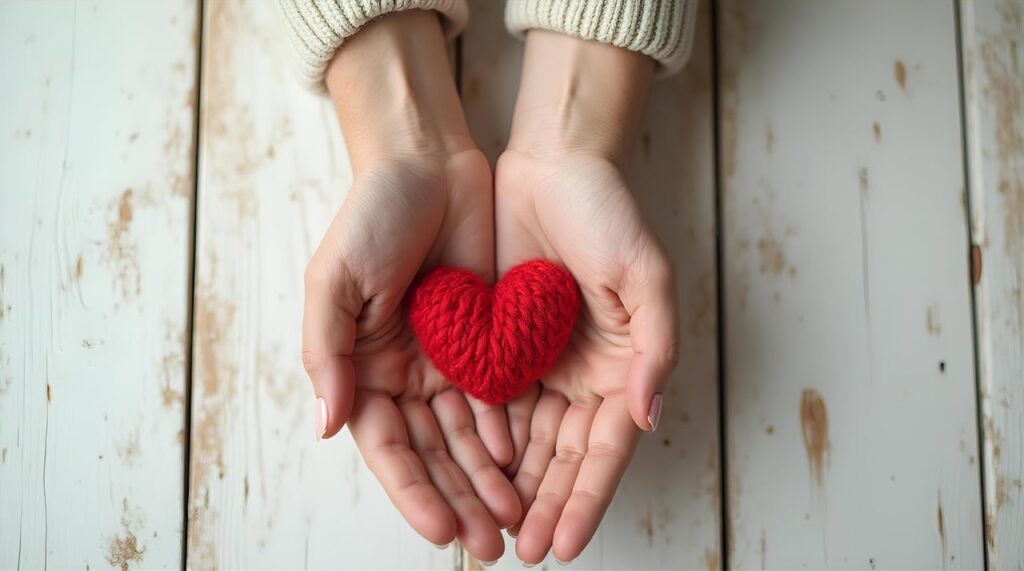 「両手のひらに赤い毛糸のハートを包み込む写真。思いやり・寄り添いの象徴。」