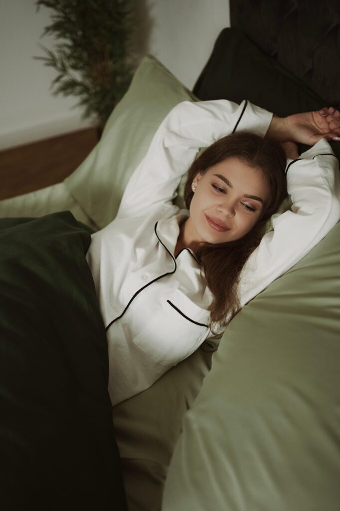 白いパジャマの女性がベッドの上で背伸びをしながら眠りにつく準備をしている様子