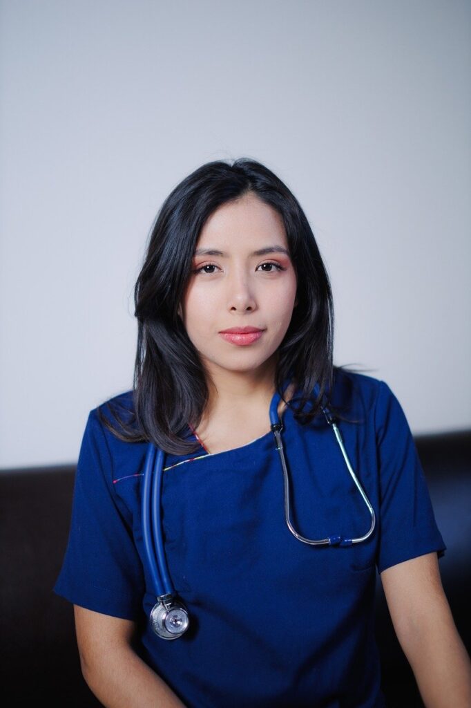 青い制服を着て聴診器を肩にかけ、正面を向いて微笑む女性看護師。訪問看護の現場を象徴するイメージ。