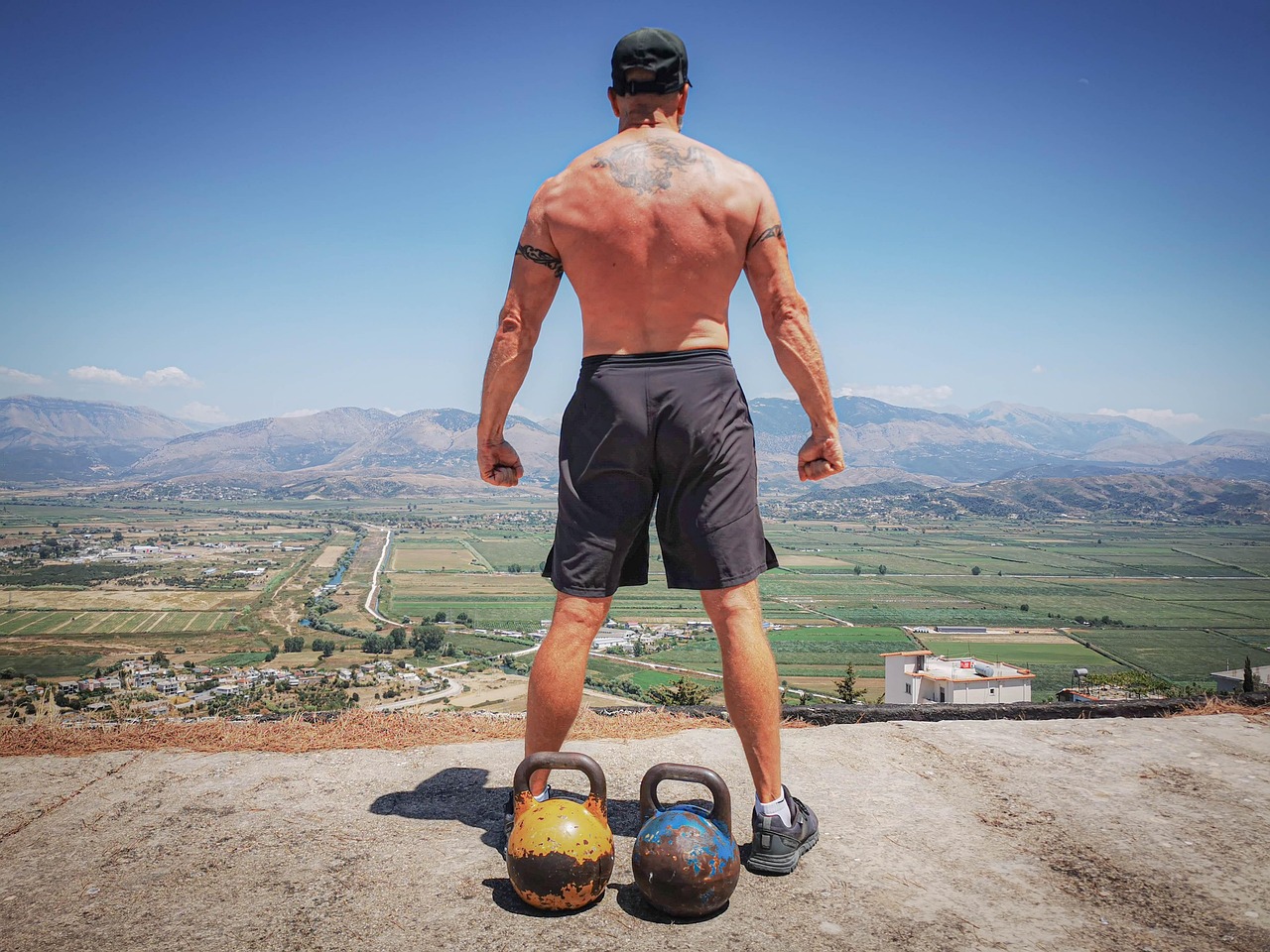 「丘の上で街を見下ろす上半身裸の男性。ケトルベルを足元に置き、筋トレを通して自分を鍛える姿を背中越しに捉えた写真。」