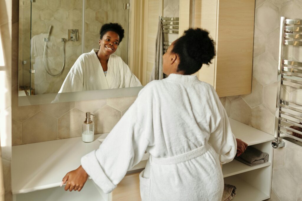 バスローブ姿の女性が鏡に向かって笑顔を見せる様子。朝のアファメーションで自分に優しく語りかけている。