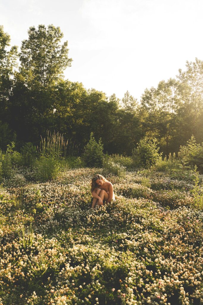 朝日が差し込む草原やお花畑の中で、女性が座りながら穏やかな時間を過ごしている様子。自然の豊かさと心の安らぎを象徴するシーン。