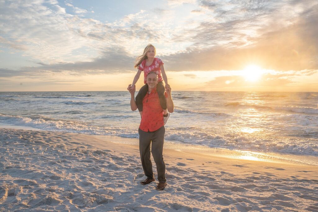 夕焼けの浜辺で父親が娘を肩車し、2人がこちらを向いて微笑む写真。家族の絆と安心感を象徴する温かなシーン。