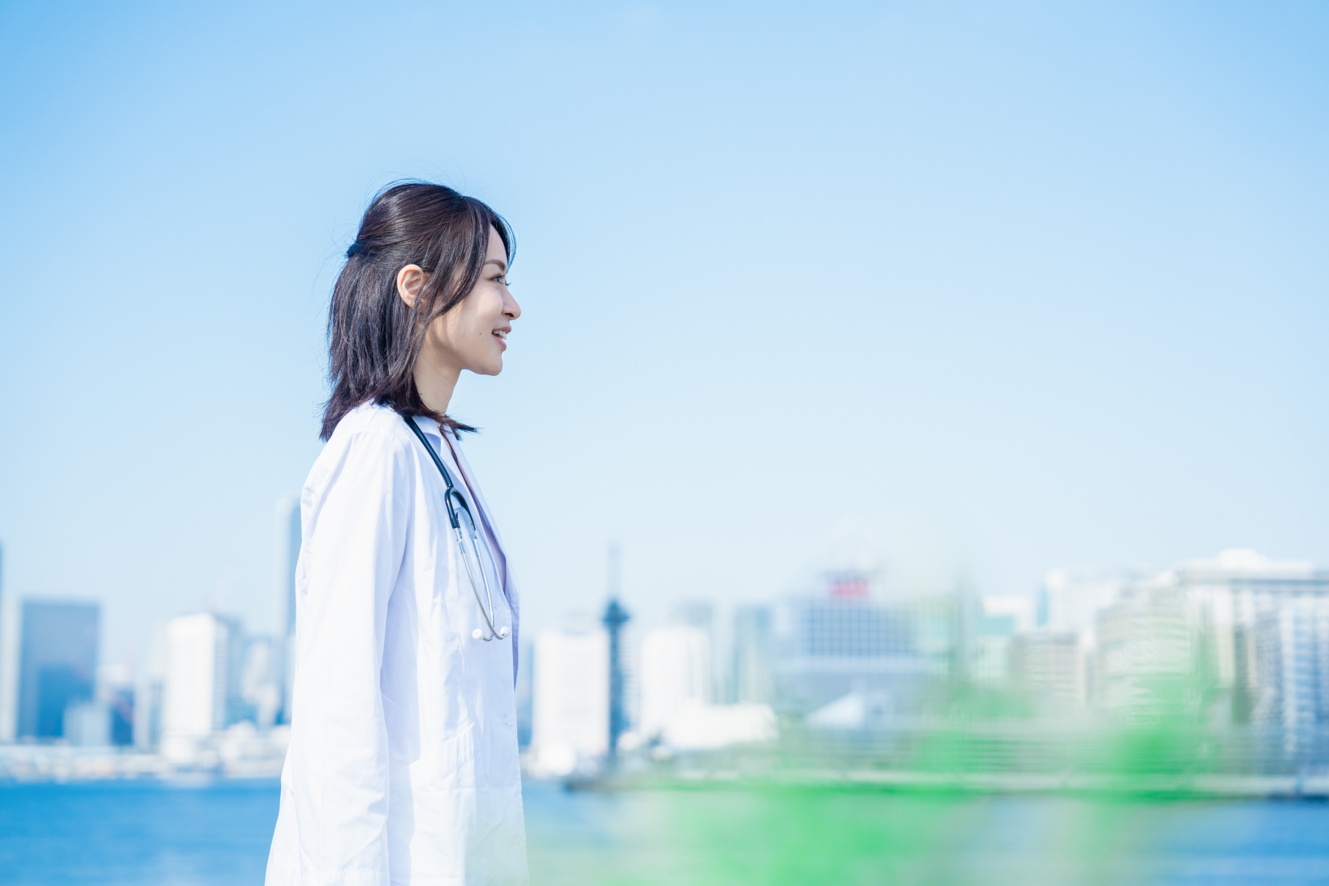 青空と海を背景に、白衣を着た女性医療従事者が横顔で前を見つめている様子。