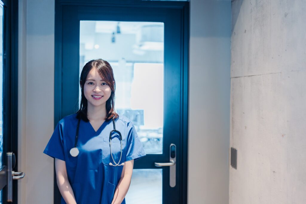 青い看護師制服を着た女性が、名札を首から下げ、ナースステーション前で笑顔を向けて立っている写真。