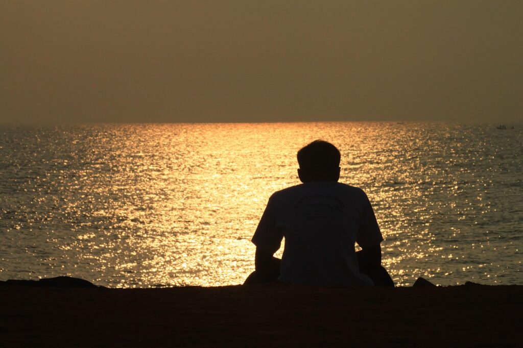 夕暮れの海を前に、ひとり座って水面を見つめる人物の後ろ姿。静かな時間が流れている様子。