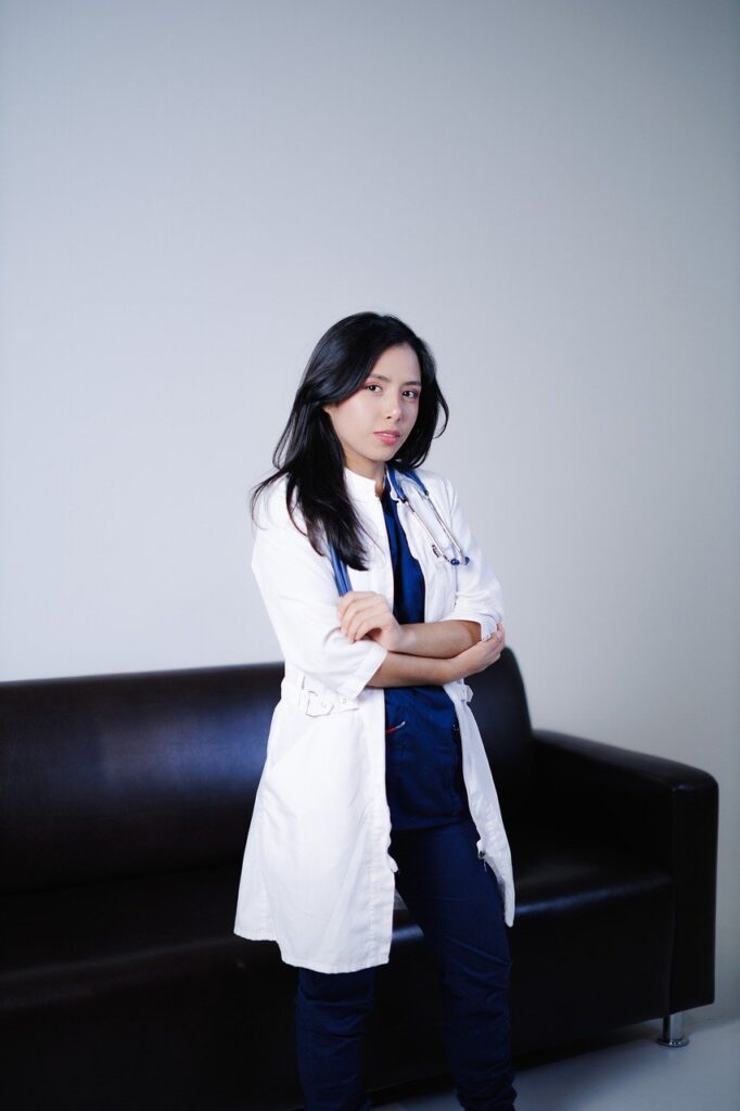 白衣を着たベテラン看護師が聴診器を首にかけ、黒いソファーに座ってカメラ目線で微笑む。背景は白い壁でシンプル。落ち着きとメンタルの安定を感じさせる写真。