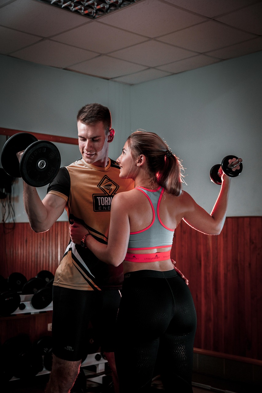 ジムで男女が向かい合い、ダンベルカールを行っている様子。楽しそうな雰囲気でトレーニングを続けるイメージを表す写真。