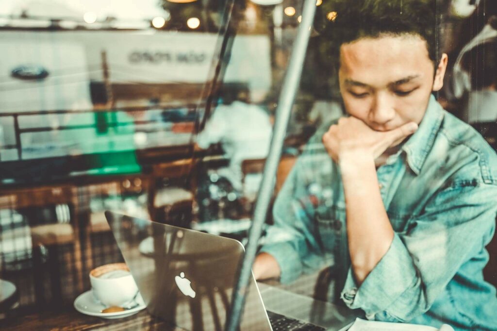 カフェでノートパソコンを前に、顎に手を当てて考え込む人物。静かな時間の中で、自分と向き合っている様子。