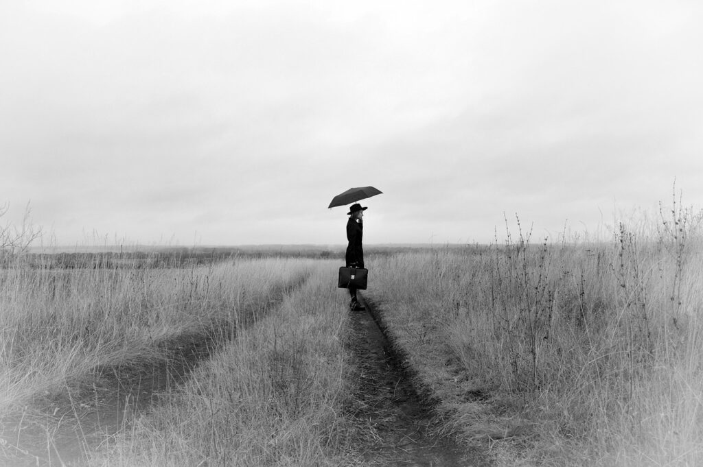 草原の一本道に立ち、傘と鞄を持つ人物の後ろ姿。真面目に生きてきた人が自分を後回しにして歩いてきた人生を象徴する風景。