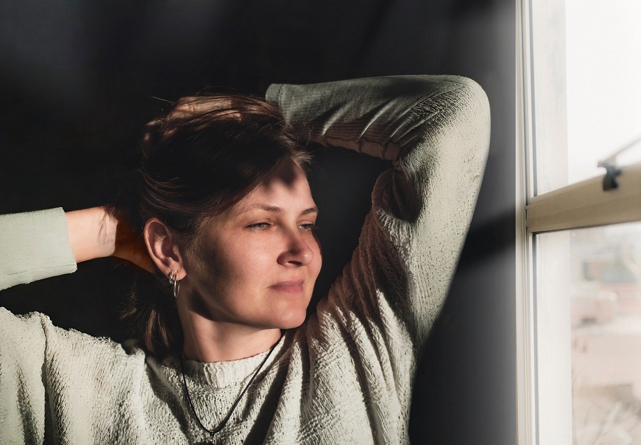 窓辺で腕を上げ、静かに外を見つめながら一息つく女性。頑張り続けてきた人の心の休息を表すイメージ。