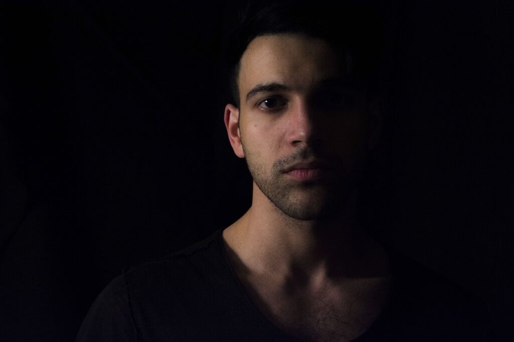 暗い背景の中で半分だけ光に照らされた男性の顔。自分を疑う心と内面の葛藤を象徴するイメージ