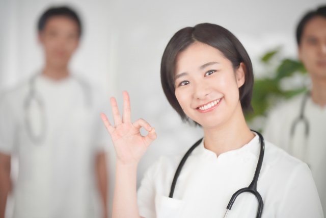 笑顔でOKサインを出す女性看護師。背景に医療スタッフがぼかして写っている。