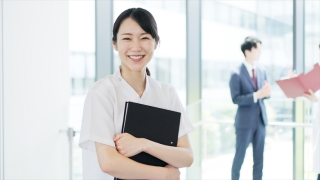 笑顔でファイルを持つ看護師と、背景にスーツ姿の人物がいる病院内の風景