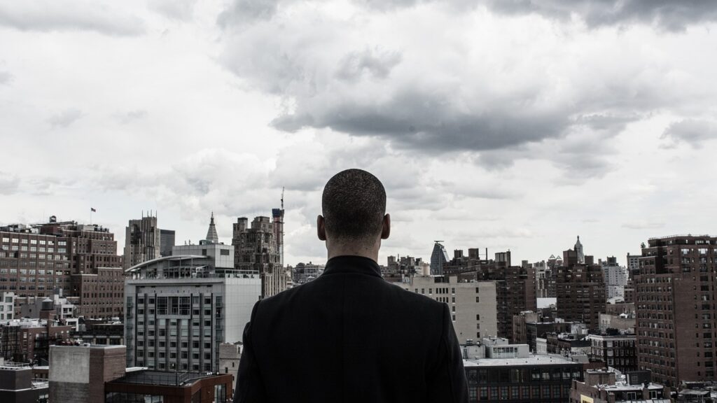曇り空の都市を背に、ビル群を見つめる男性の後ろ姿。