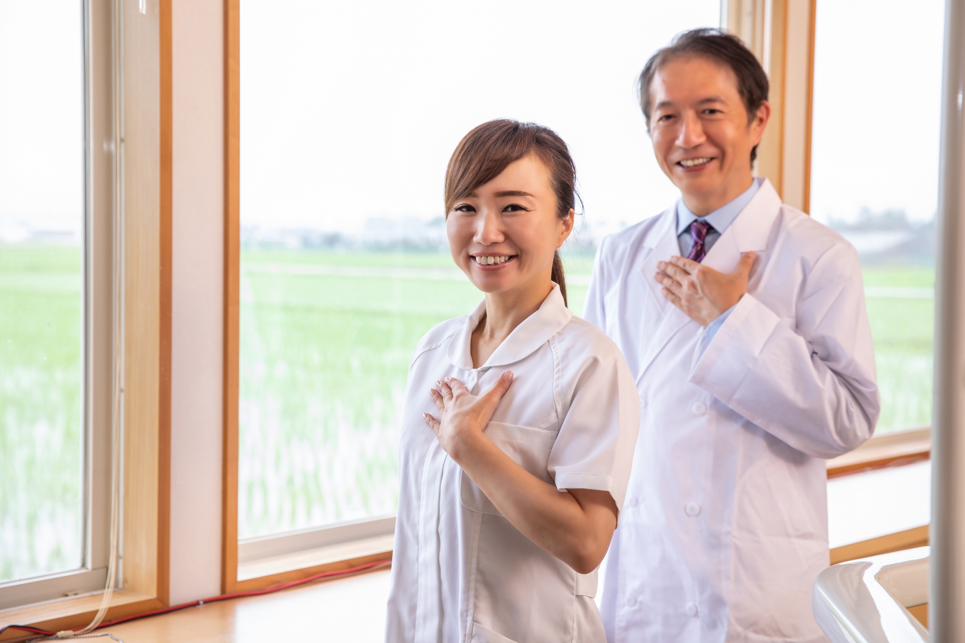 訪問看護師と医師が窓辺で微笑みながら胸に手を当てている様子